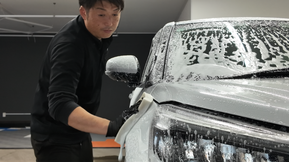 A man in a black shirt washing a white car, covered in soap suds, inside a garage.