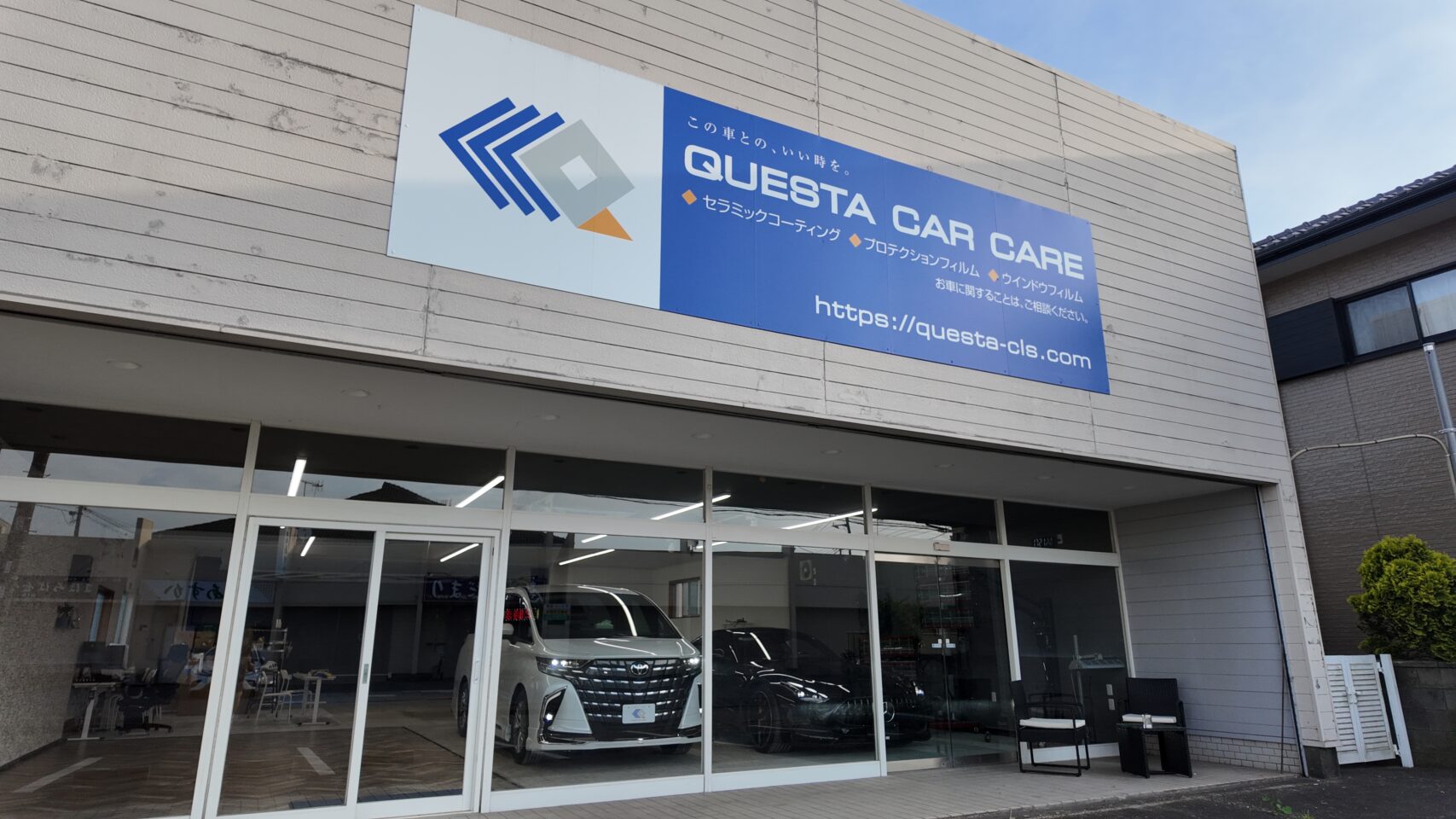 Sign on a building reading 'QUESTA CAR CARE' with a blue panel; showroom windows below show a white car inside and part of a service center exterior.