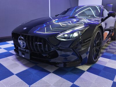 - QUESTA CAR CARE 千葉県君津市のコーティングプロショップ Black Mercedes-AMG sports car in a showroom, reflecting blue neon lights on its hood and windshield on a blue-and-white checkered floor.