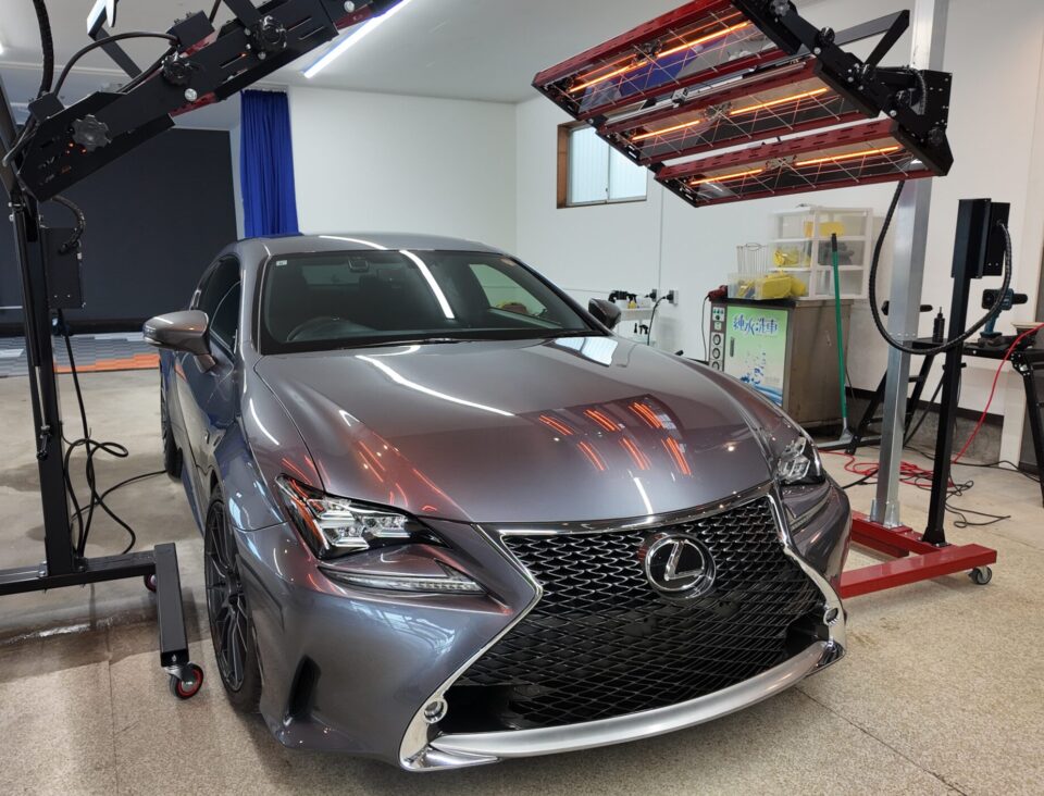 Gray Lexus sedan in a workshop surrounded by large infrared heating panels on a movable frame.