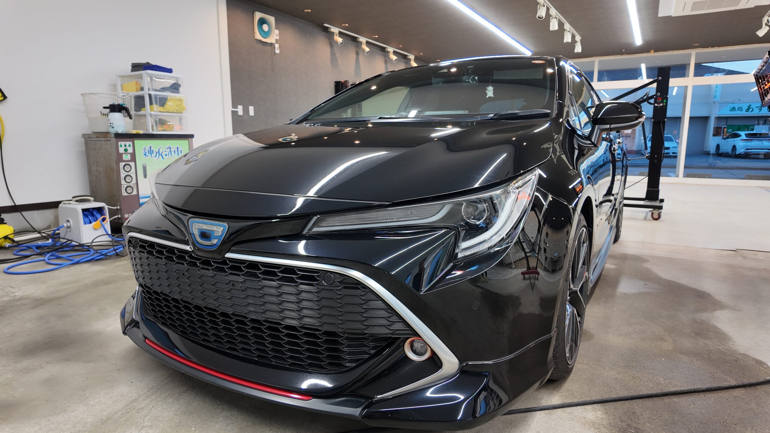 Front view of a sleek black SUV in a detailing shop, with a bold grille and headlight, hoses and equipment visible in the background.