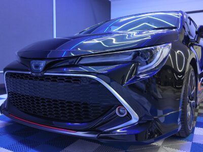 Close-up front-left view of a glossy black sedan in a blue-lit showroom with a checkered floor.