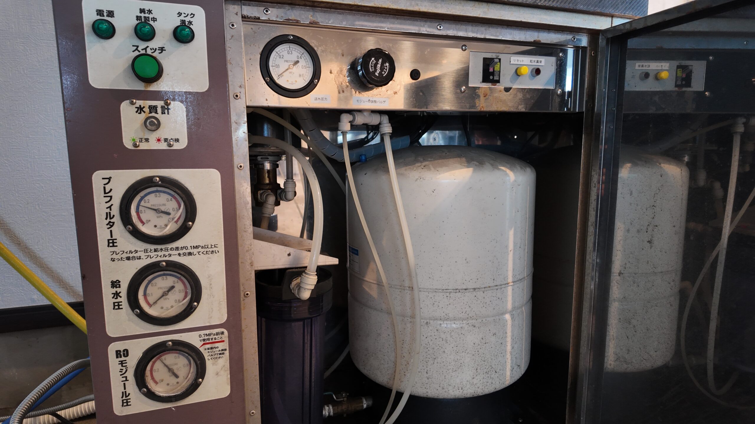 Industrial control panel with gauges and hoses beside white pressure tanks near a metal cabinet.